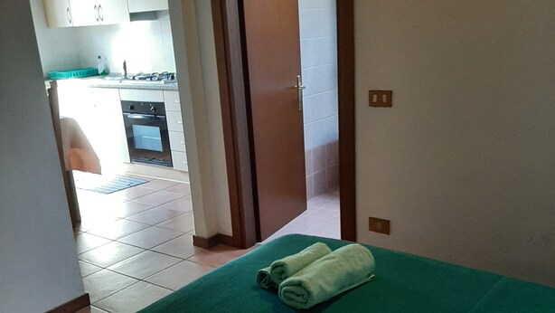 Ein Zimmer mit einer grünen Bettdecke, zwei aufgerollten Handtüchern und Blick auf eine Küchenzeile mit Schränken und Spüle.