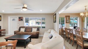 A spacious living room with a brown sofa, a white sofa, a dining table with chairs, and a ceiling fan.