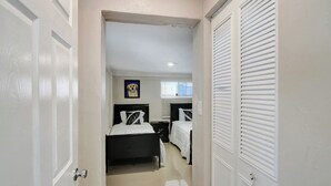 A narrow hallway leading to a bedroom with a bed, bedside tables, and a window with blinds.