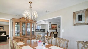 A dining room with a wooden table, chairs, a chandelier, and a kitchen in the background.