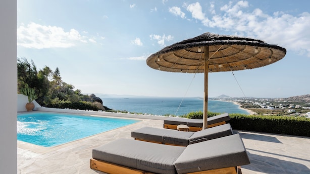 Área à beira da piscina com espreguiçadeiras e um guarda-sol de palha, com vista para a costa.