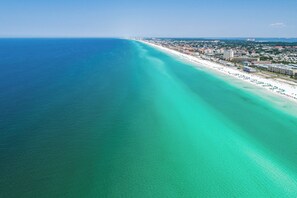 An aerial view of a coastal area with a beach, buildings, and a clear blue sea.