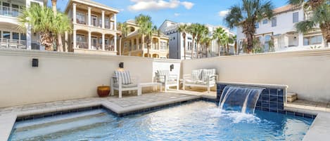 A swimming pool with a waterfall feature, surrounded by palm trees and white lounge chairs.