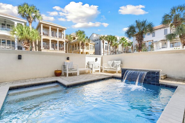A swimming pool with a waterfall feature, surrounded by palm trees and white lounge chairs.