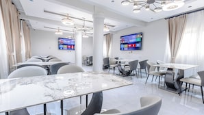 A modern dining area with marble tables and chairs, large windows with curtains, and a flat-screen TV on the wall. - Capital 11 Hotel (North Legon)