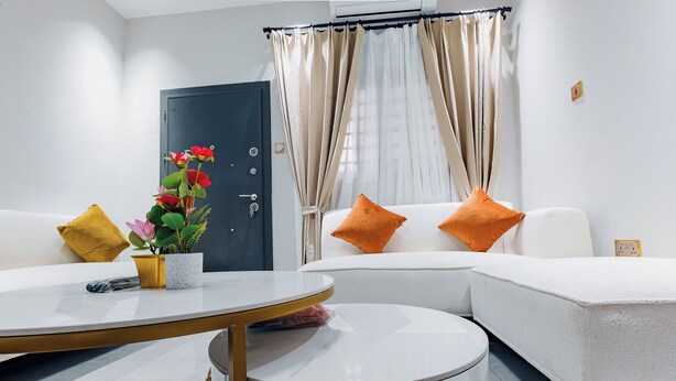 A modern hotel room with a white sofa, two orange pillows, a small round table, and a potted plant with red flowers. - Capital 11 Hotel (North Legon)