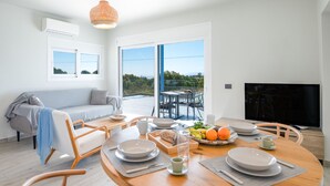 A modern living room with a sofa, dining table set for two, and a TV. Large windows offer a view of the outdoors. - Villa Antreas Mentoros (Kefalonia)
