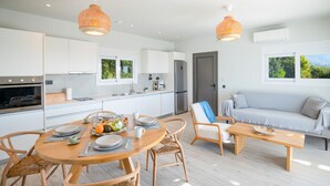 A modern kitchen with white cabinets, a dining area with a round wooden table, and a sofa with a view of greenery. - Villa Antreas Mentoros (Kefalonia)