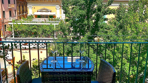 A balcony with a wicker table and two chairs, overlooking a cityscape with buildings and trees. - Third floor (Pisa)