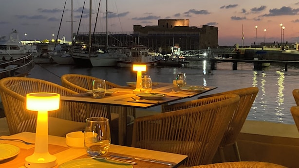 A waterfront dining area with tables set for two, overlooking a marina with boats and a historic building. - Luxury 3 Bedroom Penthouse apartment with sea view.
10 mins walk to Venus Beach (Paphos)