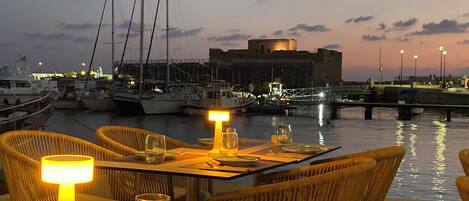 Un área de comedor frente al agua con mesas dispuestas para dos, con vistas a un puerto deportivo con barcos y un edificio histórico.