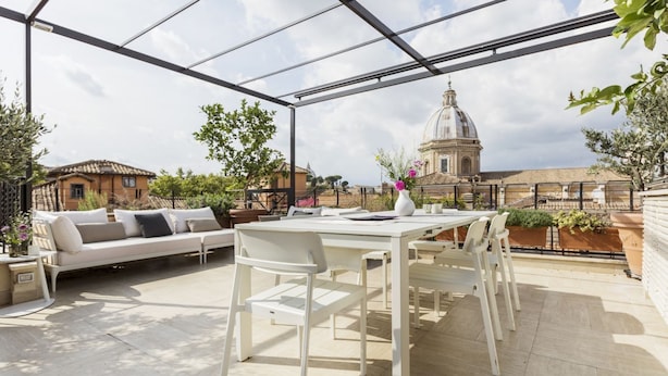 Un patio esterno con tavolo e sedie bianchi, un divano bianco con cuscini e vista su un edificio storico con una cupola.