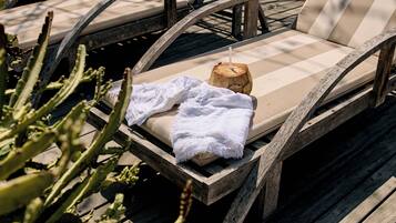 Una silla de lounge de madera con un cojín beige y una toalla blanca, un taburete de madera con una pajita en una cáscara de coco, y un cactus en primer plano.