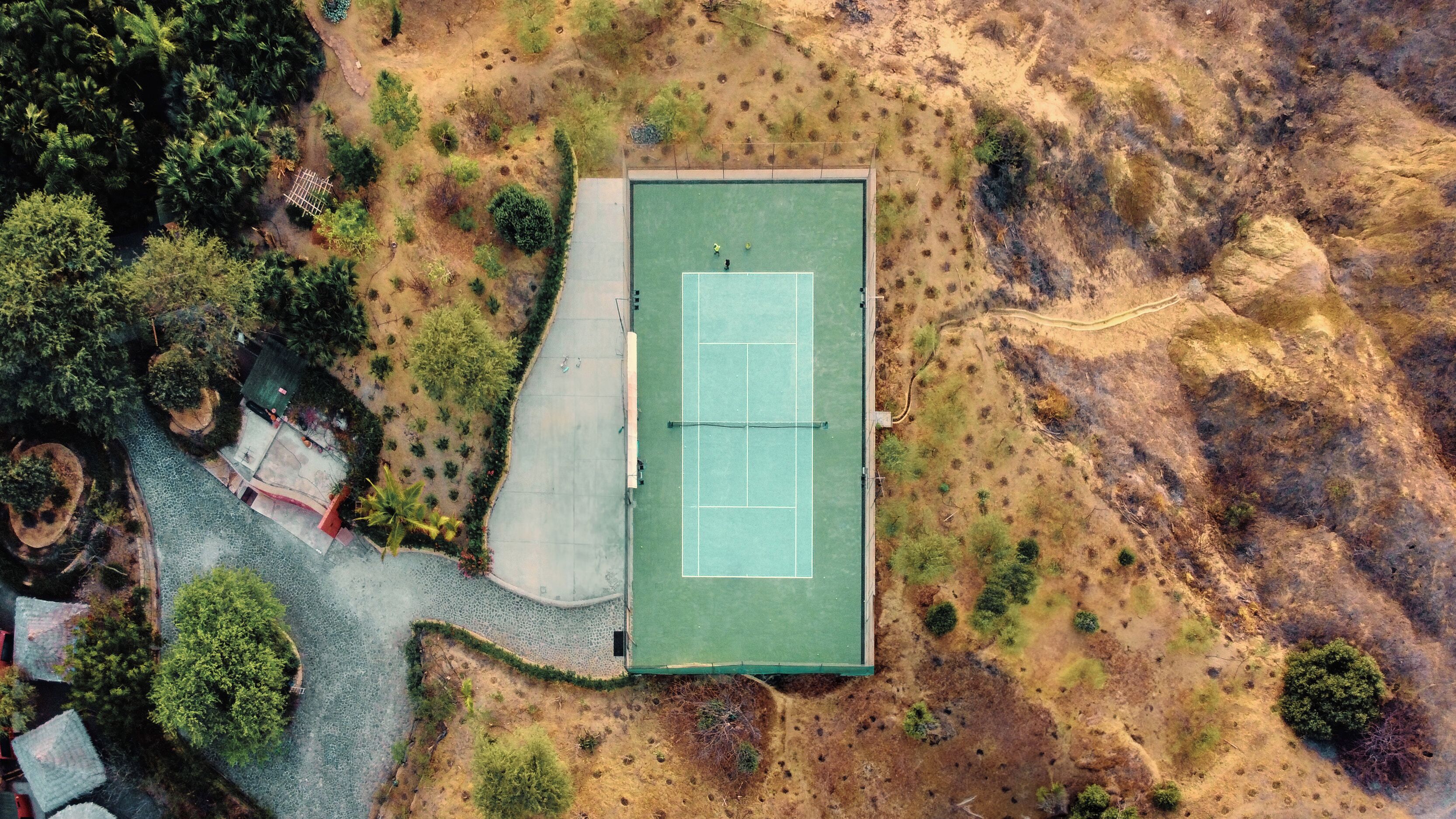 An aerial view of a tennis court surrounded by a landscaped area with trees and shrubs.