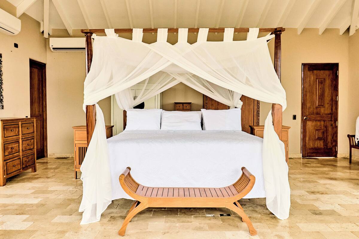 A four-poster canopy bed with a wooden bench in front, wooden furniture, and a tiled floor.