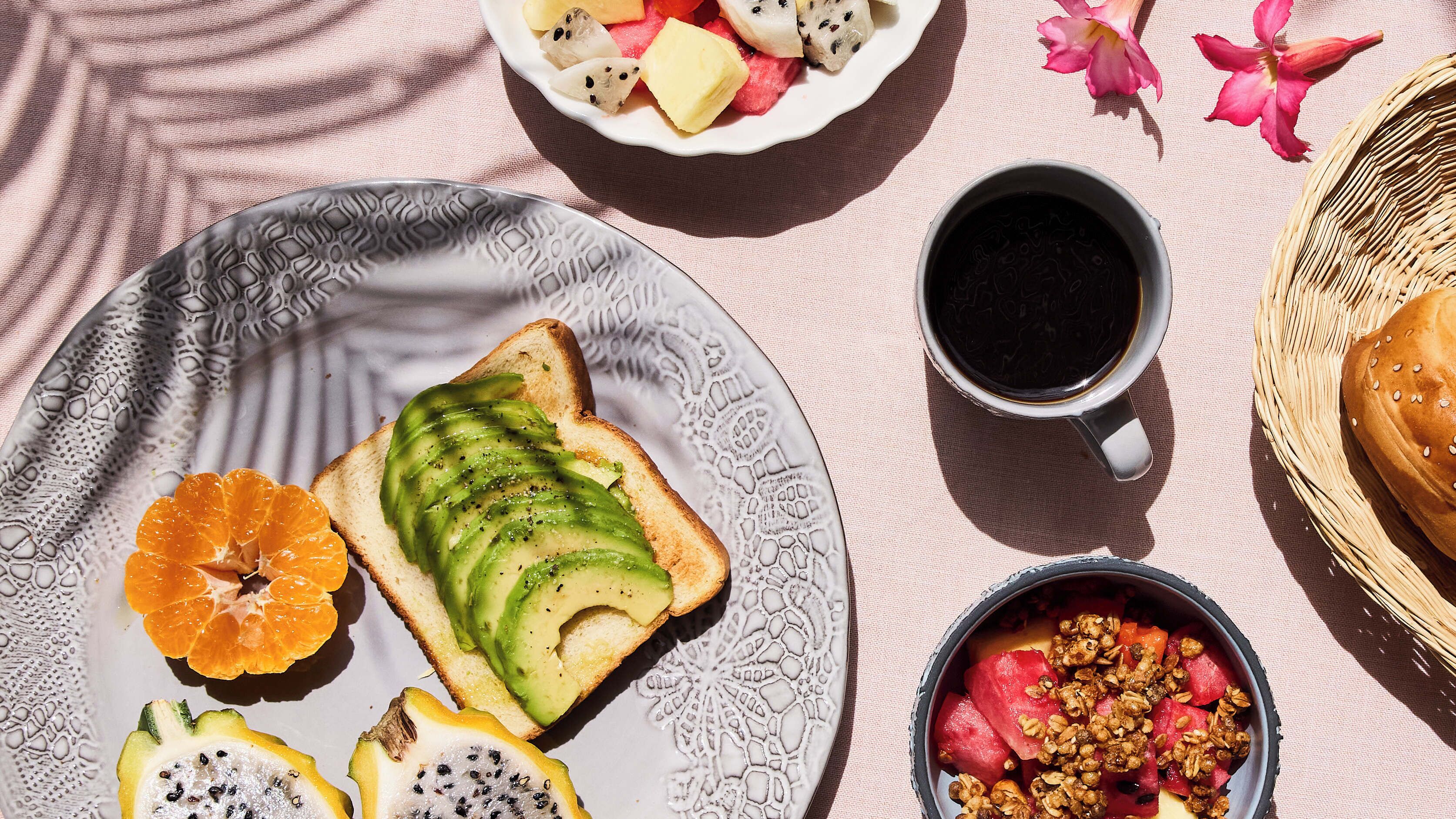 Un desayuno con tostadas cubiertas de aguacate, un tazón de fruta variada, una taza de café y una cesta de pan.