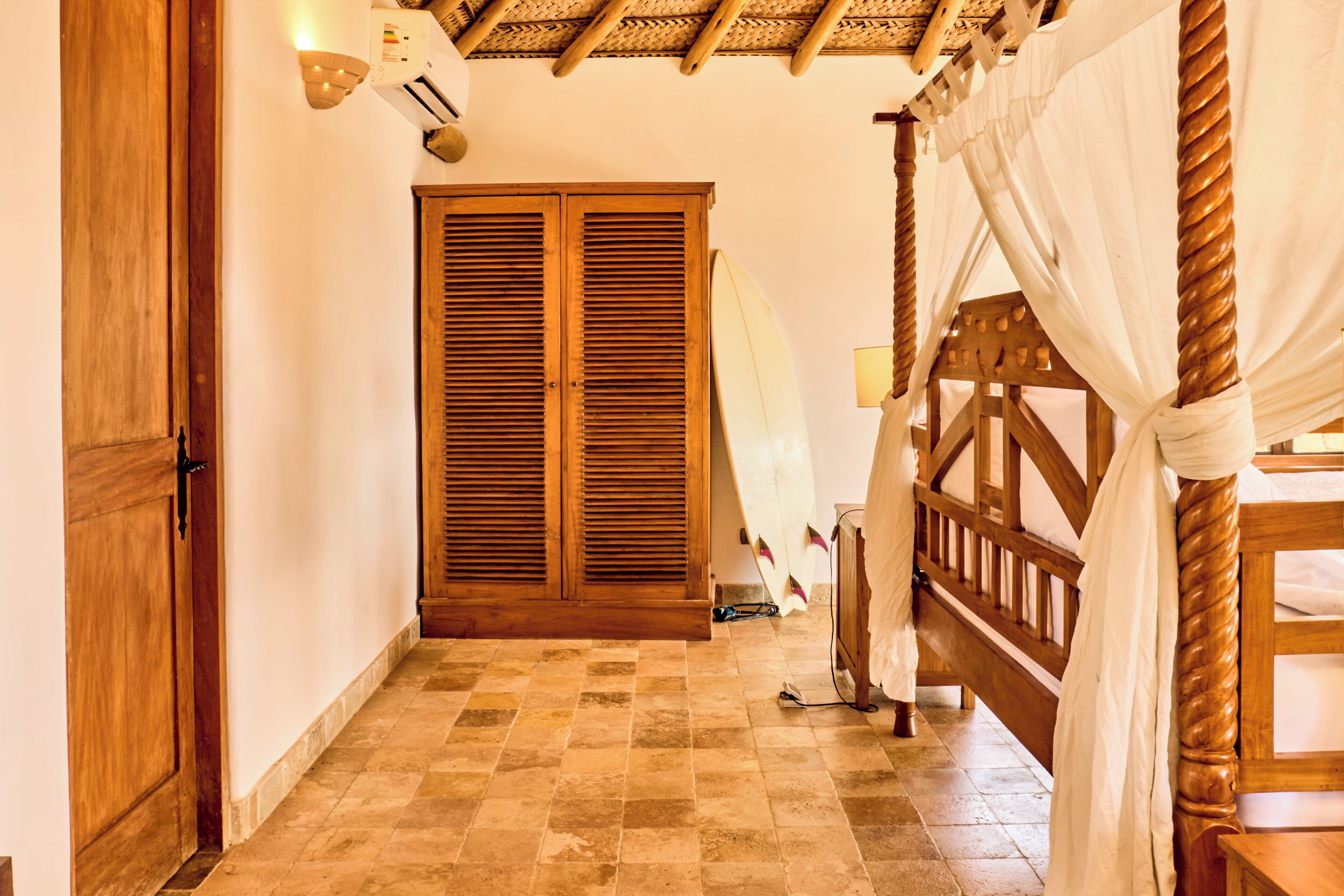 Una habitación con puerta de madera, un ventanal con contraventanas, una cama con cabecera de madera y una cortina blanca.