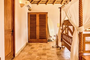 Una habitación con puerta de madera, un ventanal con contraventanas, una cama con cabecera de madera y una cortina blanca.