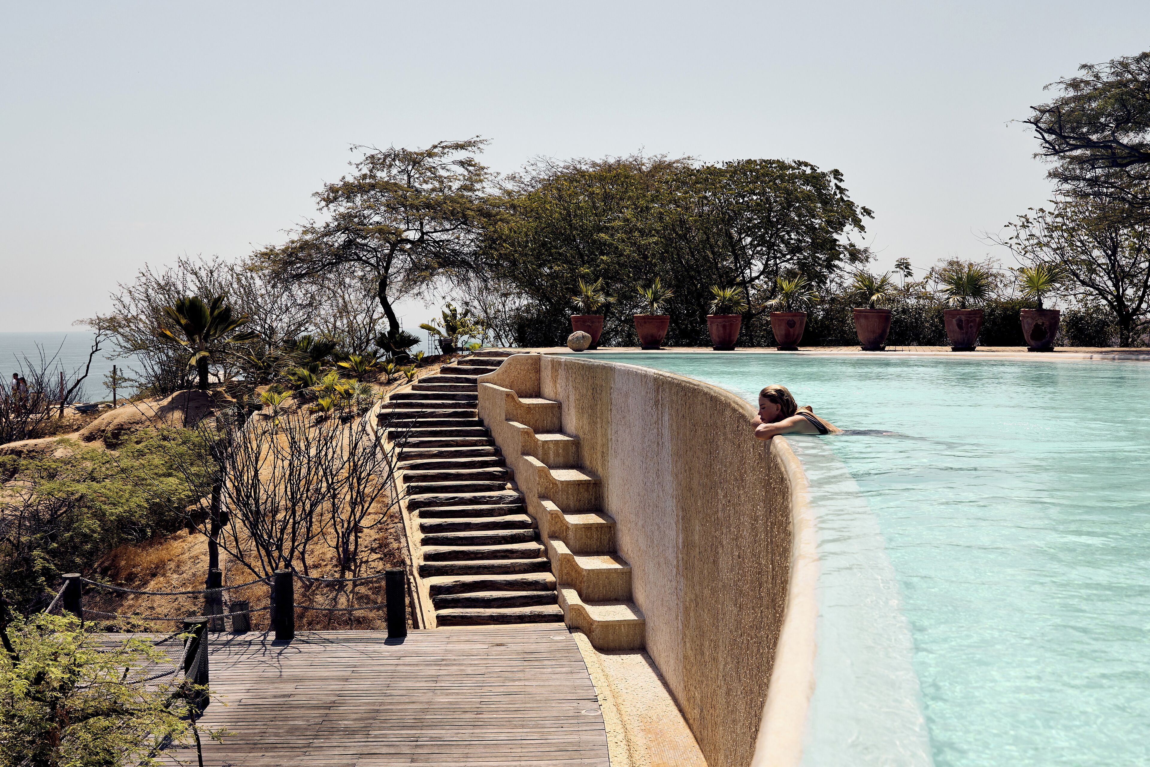 Ein Pool mit Holzterrasse und Stufen zum Wasser, umgeben von Bäumen und Topfpflanzen.