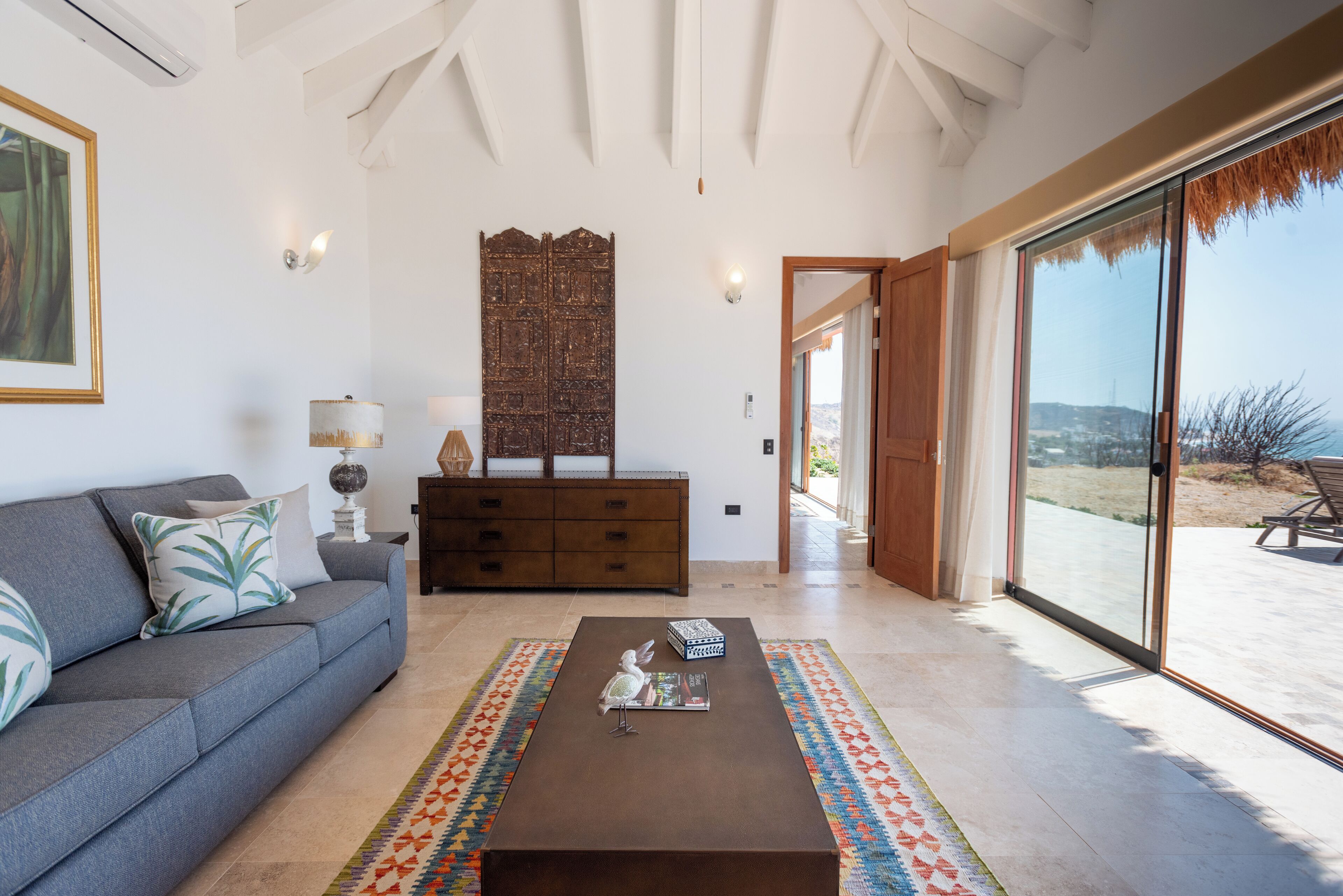 Una sala de estar con un sofá, una mesa de centro, un tapete estampado, un aparador de madera y un ventanal con vistas al exterior.