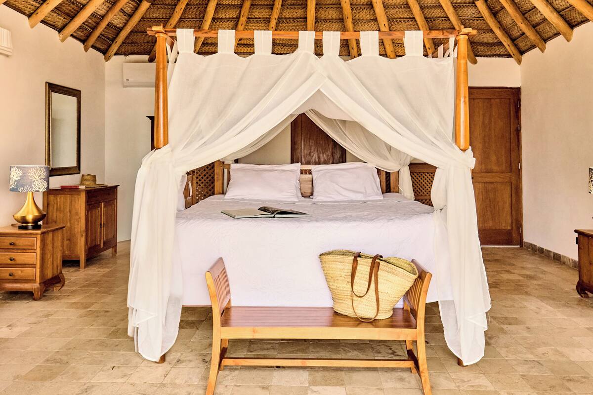 A four-poster bed with a canopy, a wooden bench, a wicker basket, and a bedside table with a lamp.