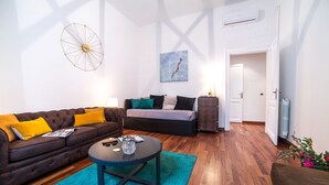 A modern living room with a brown sofa, a blue rug, and a coffee table. There is a painting on the wall and a door leading to another room.