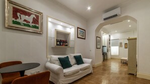 A modern living room with a white sofa, green cushions, a round table, and orange chairs. There are framed artworks on the walls and a wine rack above the sofa. - The Mirror Suite - Spanish Steps (Roma)