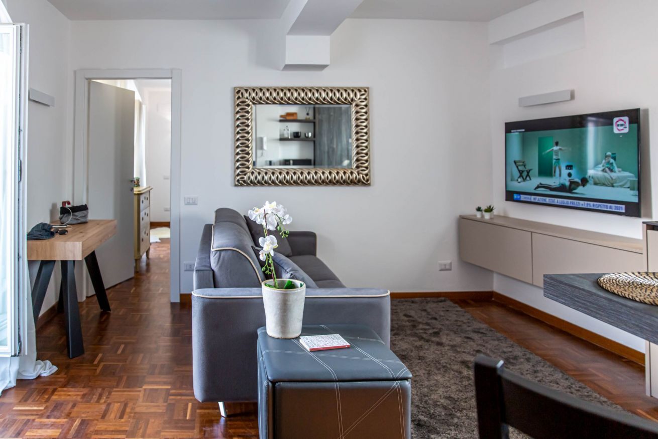 A modern living room with a grey sofa, a coffee table with a vase of flowers, a TV mounted on the wall, and a wooden floor.