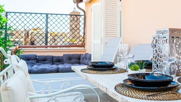 Un espacio exterior para comer con una mesa blanca dispuesta para dos, con individuales tejidos, platos y vasos. Se observa un banco acolchado azul y una silla blanca al fondo.