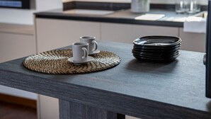 Una encimera de cocina con una pila de platos negros, dos tazas blancas y un posavasos tejido.