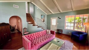 Una sala de estar con un sofá rosa, un sillón azul, una escalera y un amplio ventanal con vistas al verdor.