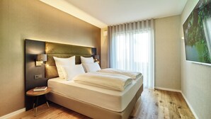 A modern hotel room with a large bed, a nightstand, a wall-mounted TV, and a large window with curtains.