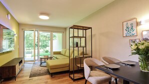 A modern living room with a sofa, a dining table with chairs, a bookshelf, and a large window with a view of greenery.