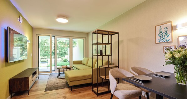 A modern living room with a sofa, a dining table with chairs, a bookshelf, and a large window with a view of greenery. - Design apartment for couples or families with a view of the countryside and garden (Trassem)