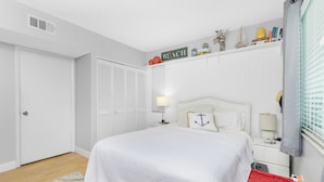 Un dormitorio con cama, mesitas de noche, un estante con libros y objetos decorativos, una ventana con persianas y una puerta.