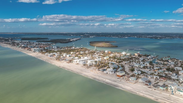 Vista aérea de un pueblo costero con playa, cielos despejados y aguas tranquilas.