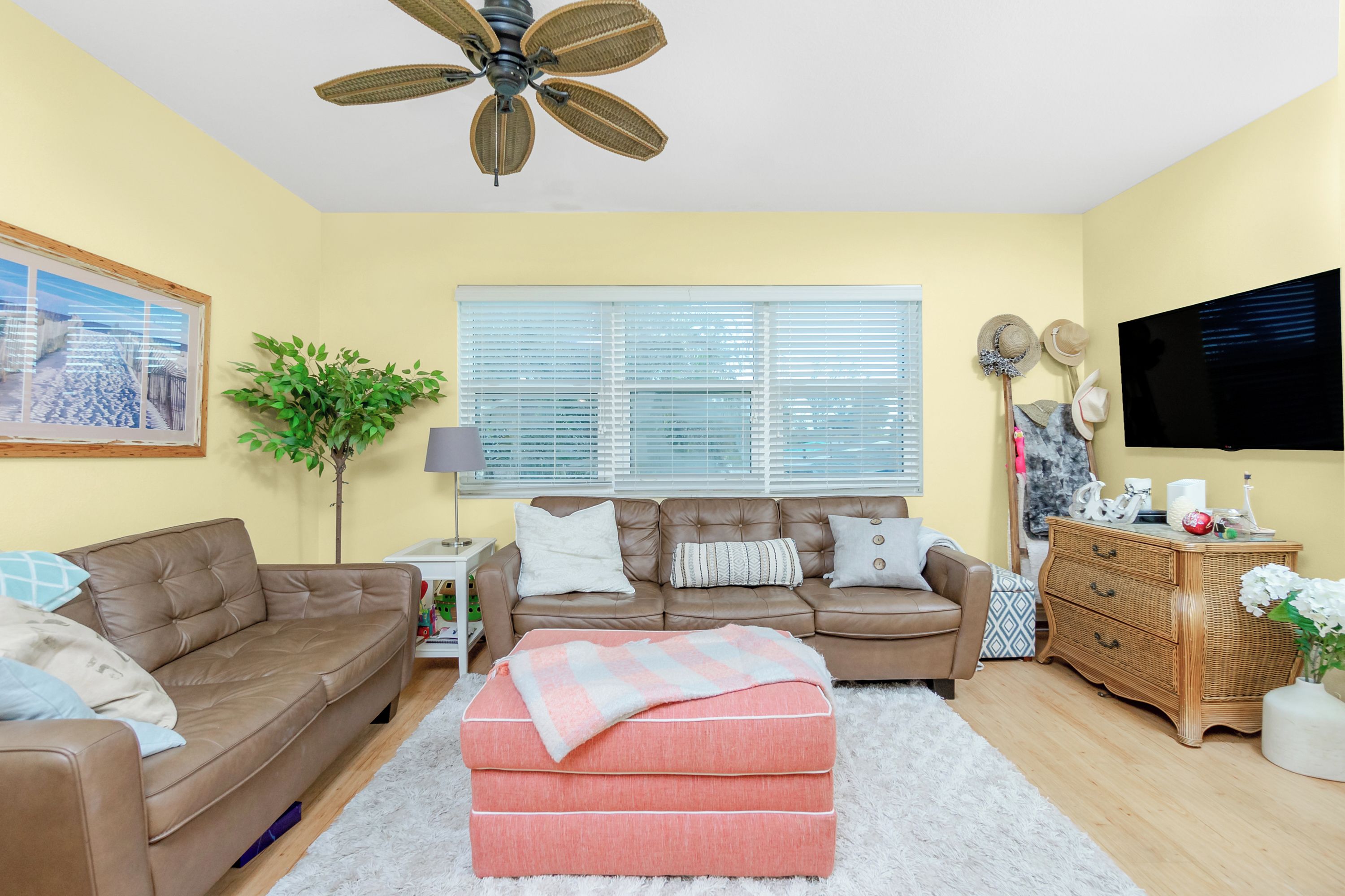 A living room with two brown leather sofas, a pink ottoman, a ceiling fan, a television, and a window with blinds.