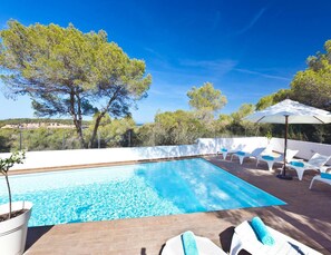 Une piscine rectangulaire à l’eau d’un bleu limpide, entourée de chaises longues blanches dotées de coussins bleus, et un grand parasol qui offre de l’ombre.