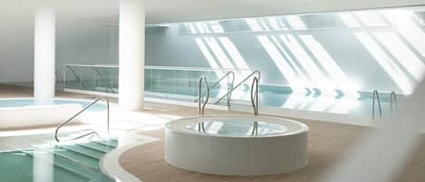 Una piscina interior moderna con un jacuzzi circular, barandillas de vidrio y suelo de madera.