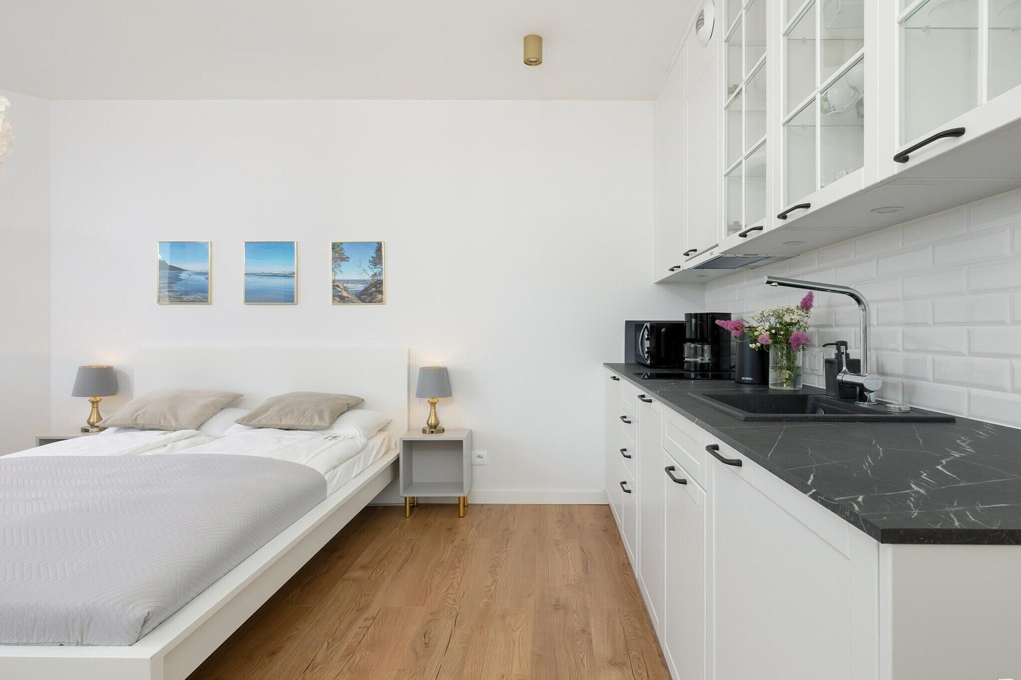 A modern bedroom with a bed, bedside lamps, and a nightstand. Adjacent is a kitchen with a black countertop, white cabinets, and a sink.