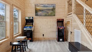 Una habitación con paredes de madera, un cuadro de un paisaje, máquinas de arcade, una mesa con sillas y una escalera con barandilla.