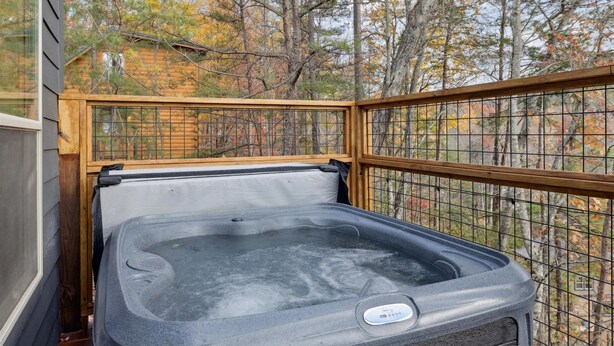 Un jacuzzi con panel de control digital, rodeado por una terraza de madera y una valla metálica, con árboles otoñales al fondo.