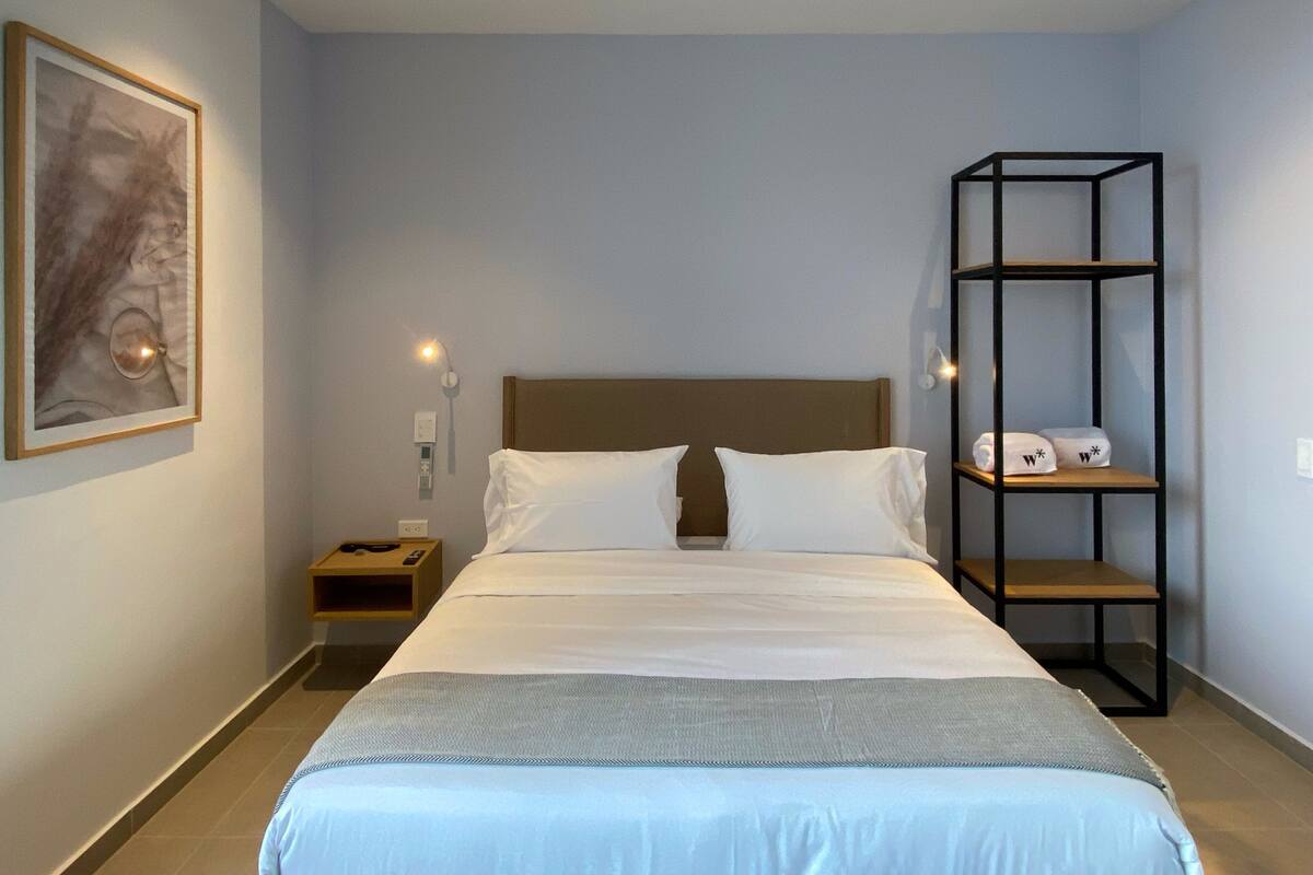 Ein ordentlich eingerichtetes Hotelzimmer mit Bett, Nachttisch, Wandregal und einem gerahmten Kunstwerk.