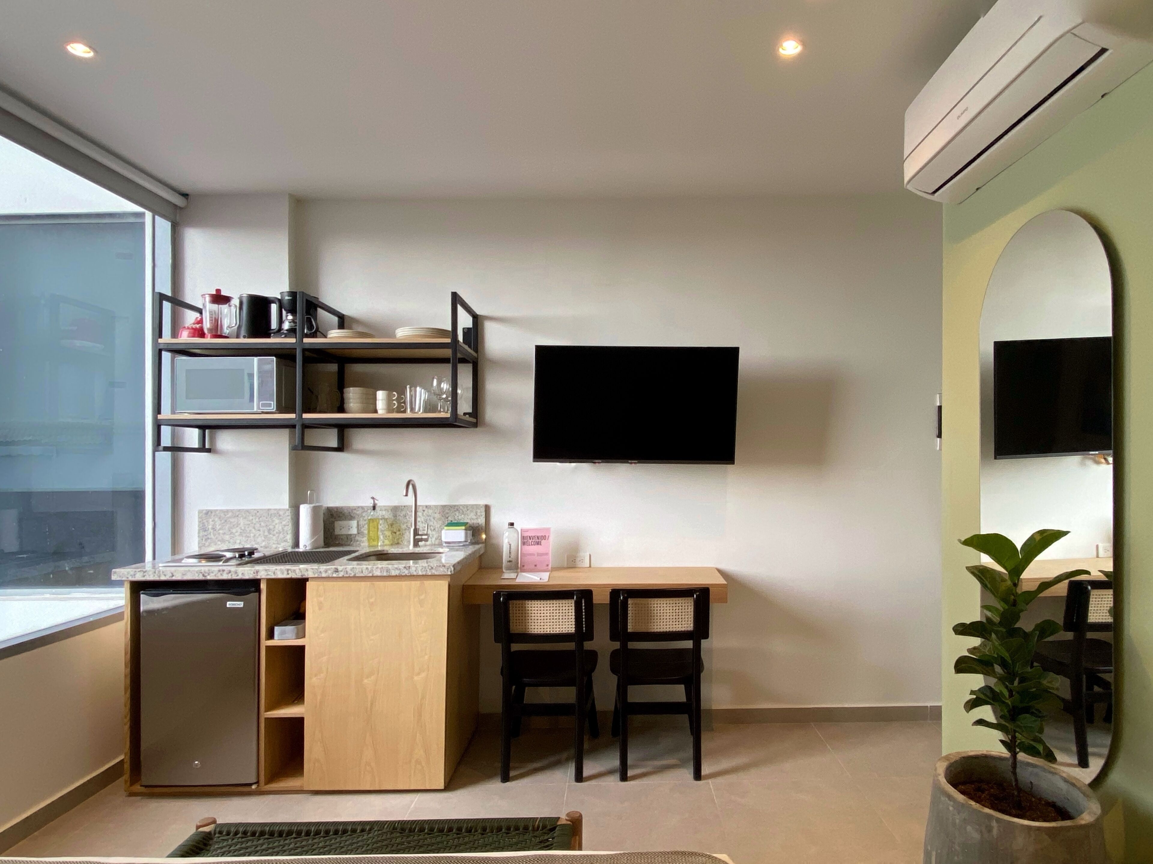 Een moderne keuken met een ingebouwde magnetron, een spoelbak en een kleine koelkast. Er staan twee stoelen en een klein tafeltje. Een flatscreen televisie is aan de muur boven het tafeltje bevestigd.