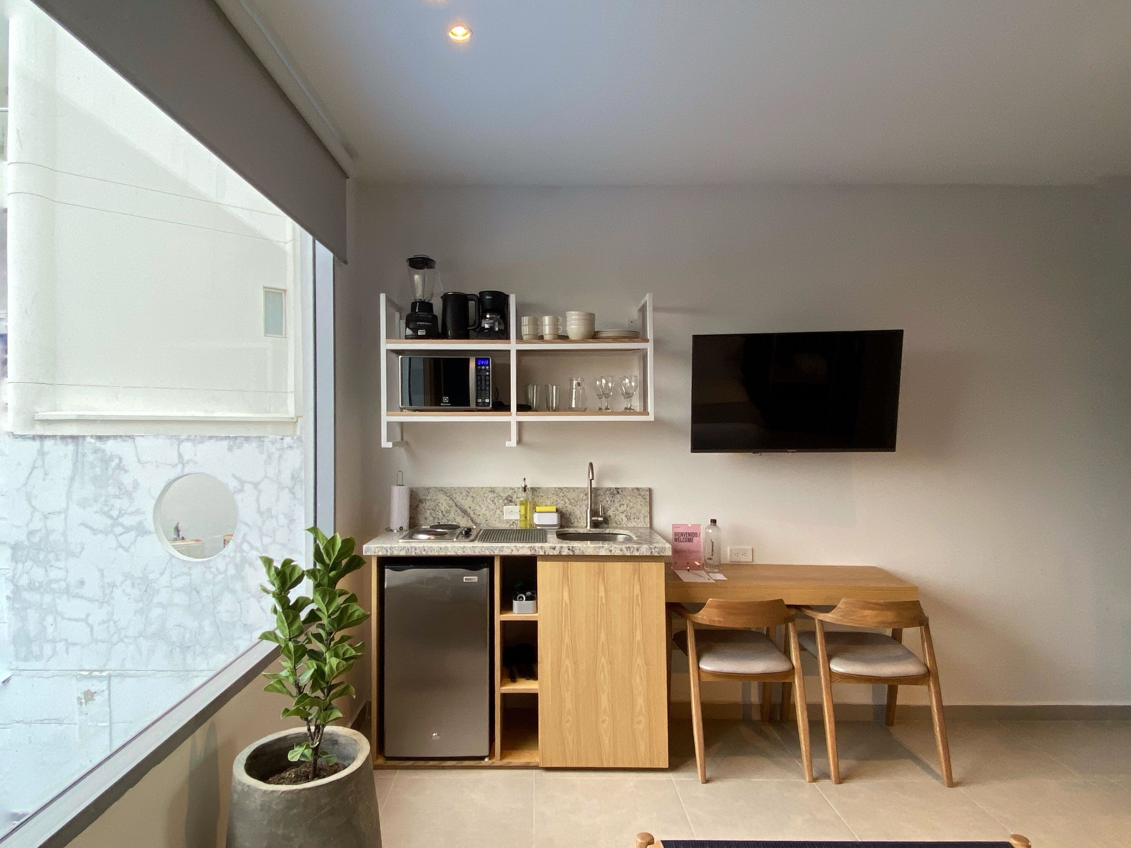 Een moderne keuken met een koelkast, magnetron en koffiezetapparaat op het aanrecht. Er is een eettafel met stoelen en een televisie aan de muur bevestigd.