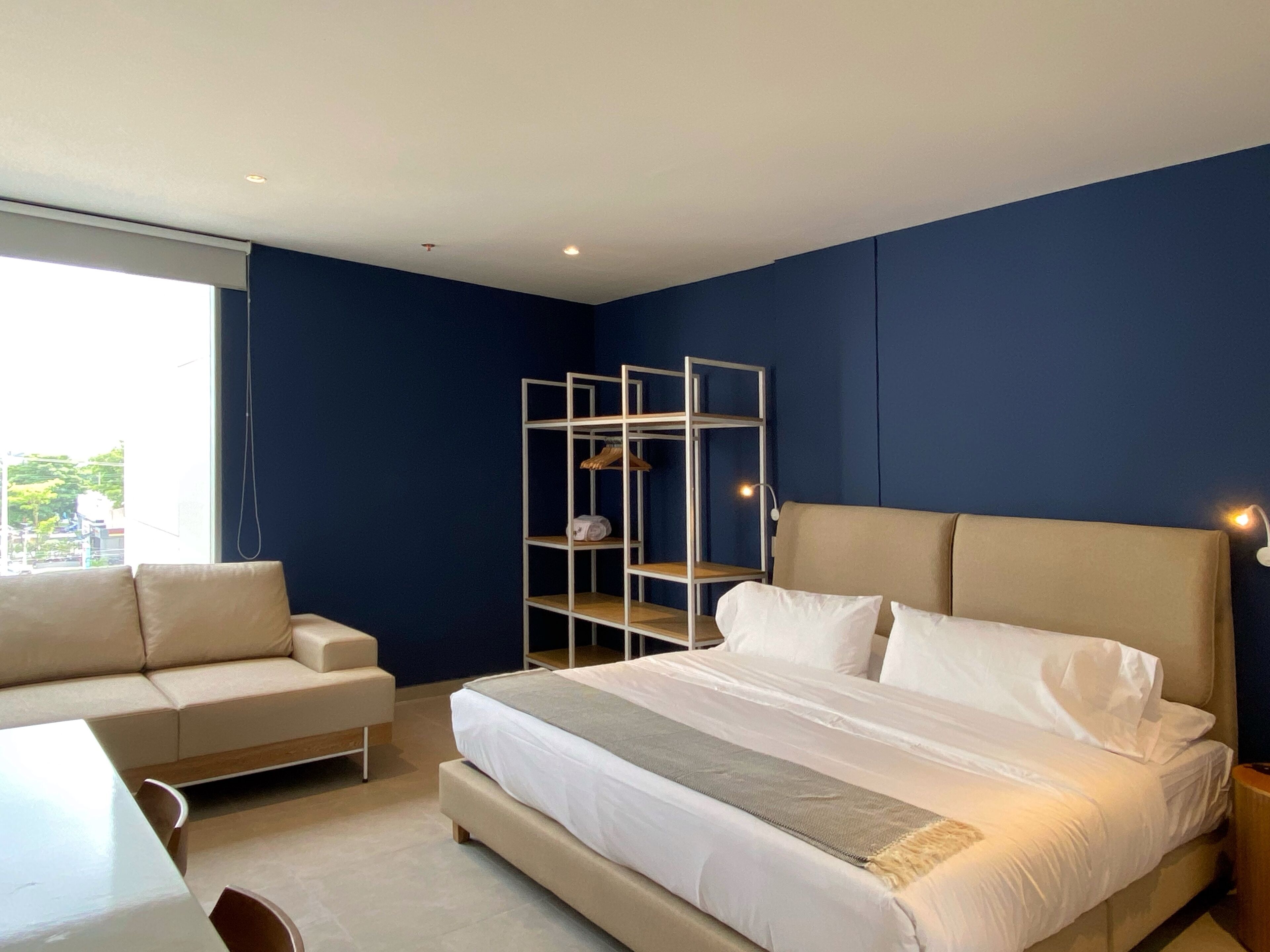 A modern bedroom with a large bed, a sofa, a metal shelving unit, and a window with a view of buildings.