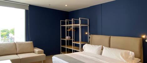 A modern bedroom with a large bed, a sofa, a metal shelving unit, and a window with a view of buildings.