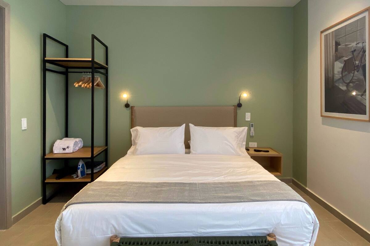 Ein Hotelzimmer mit einem Bett, einem Nachttisch, einer Ablage mit Handtuch und Bügeleisen sowie einem Wandbild.