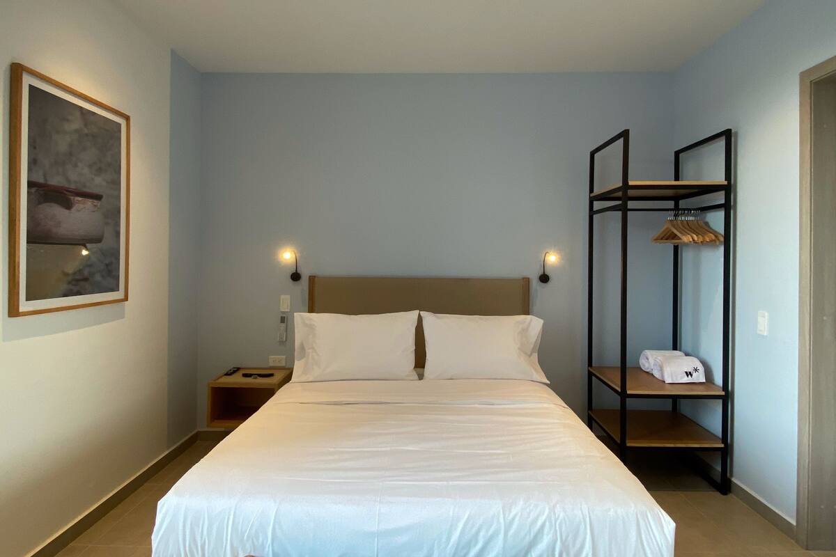 Ein Hotelzimmer mit Bett, Nachttischen, einem Wandregal mit Handtüchern und einem gerahmten Bild.