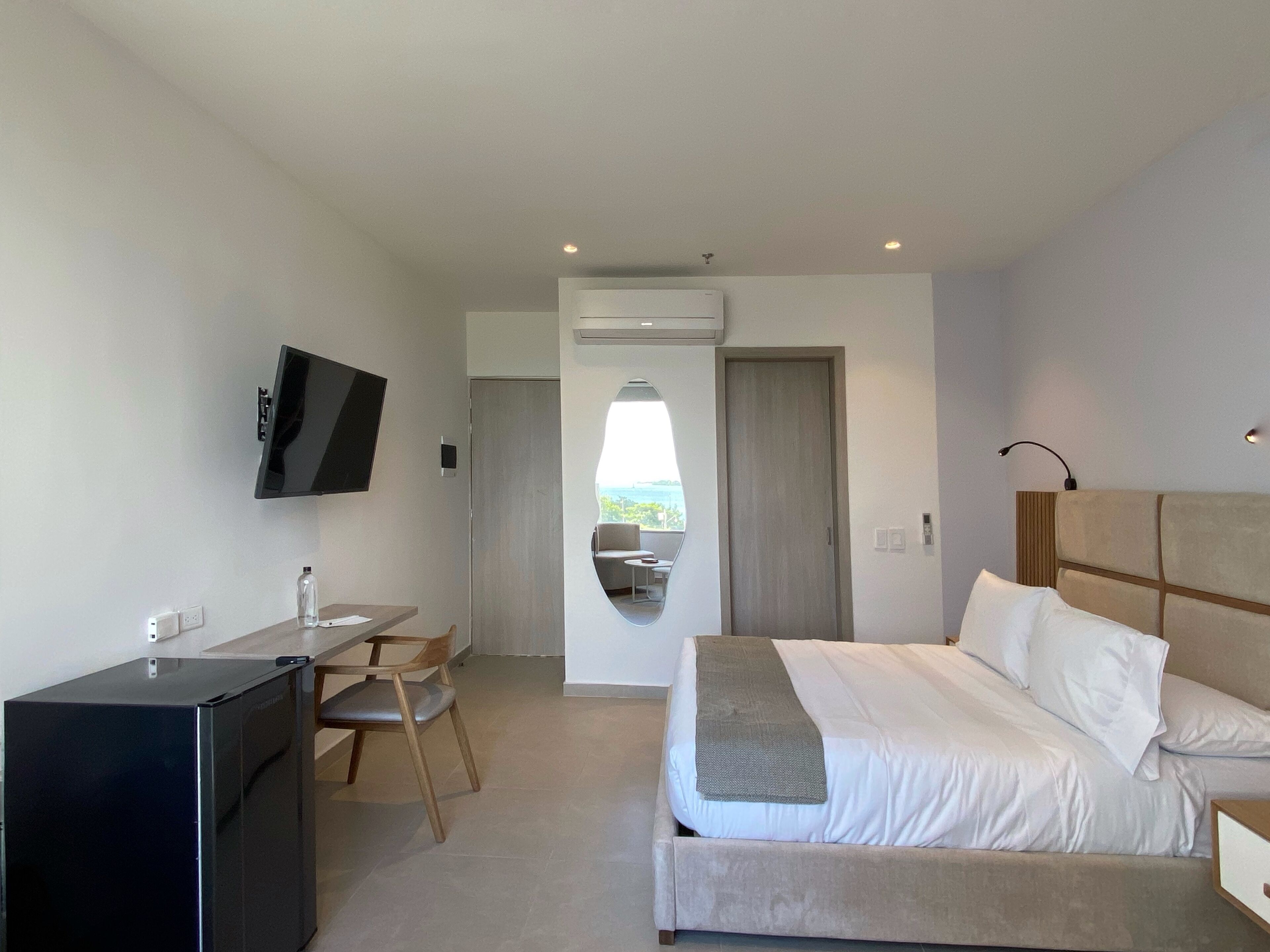 A modern hotel room with a bed, a desk, a chair, a TV, and a view through a window.